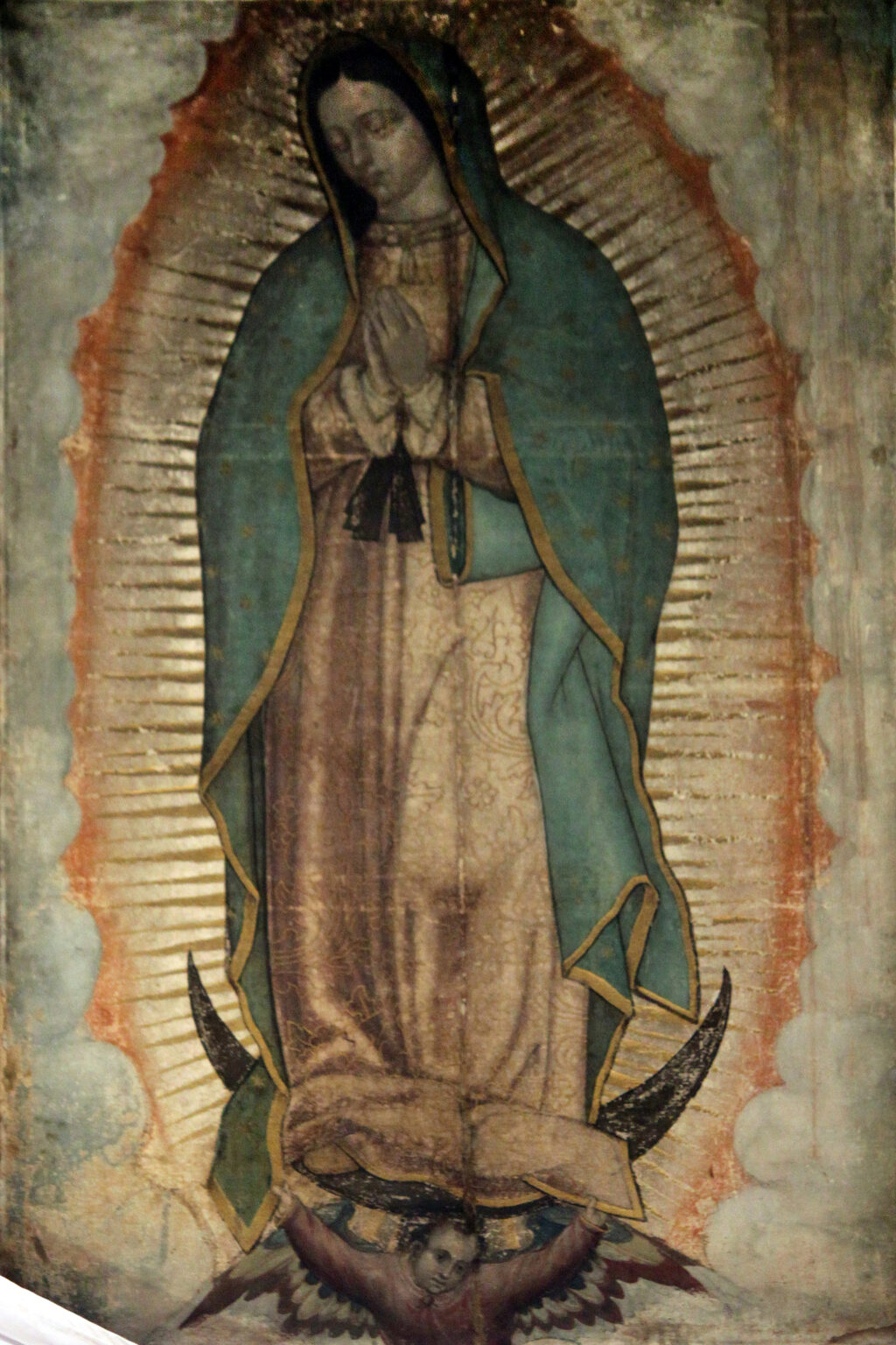 A young woman with dark hair and downcast eyes stands on a crescent moon held up by an angel, wrapped in a star covered blue green mantle, surrounded by rays of gold light, on the coarse weave of a cloak.
