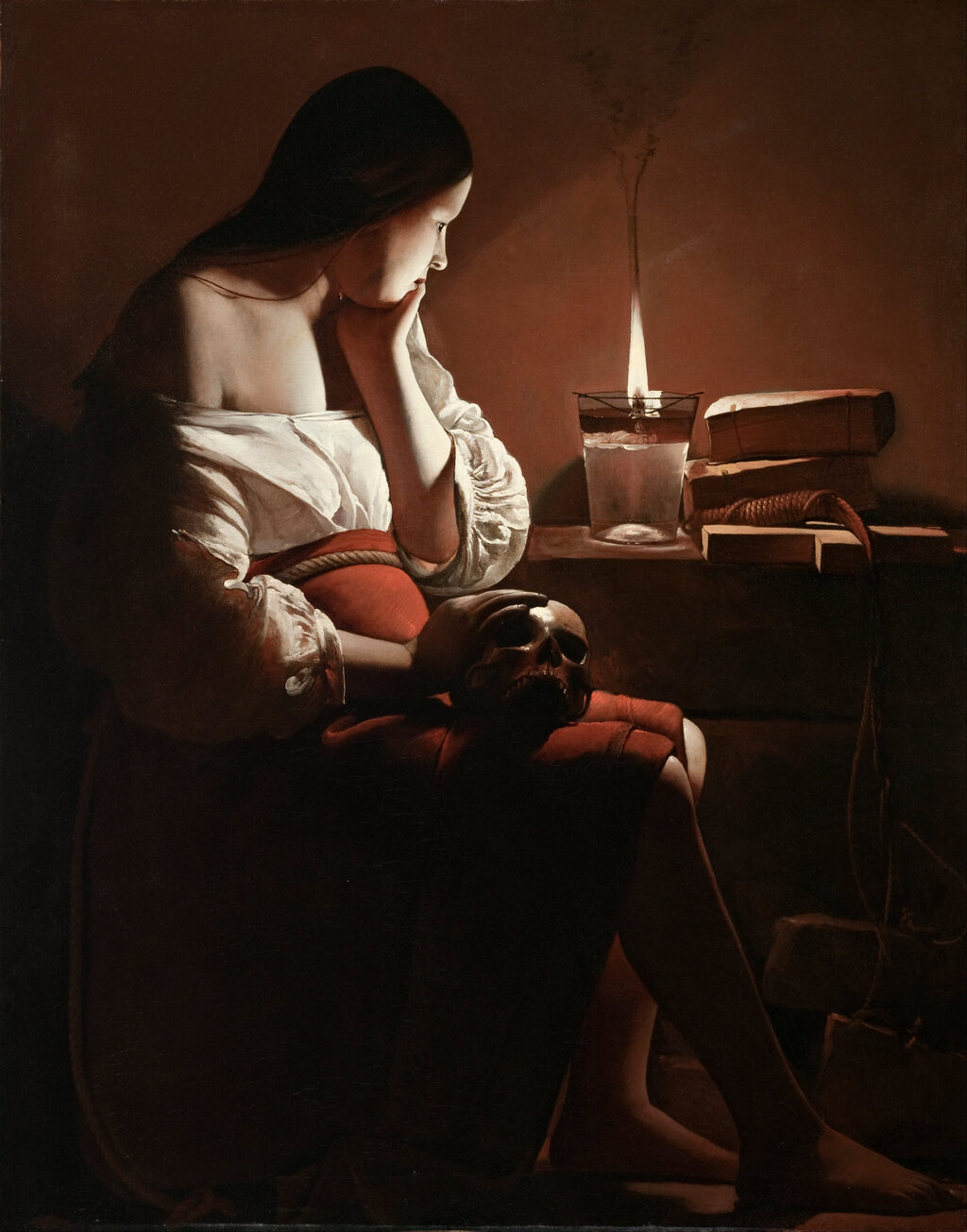 A young woman sits in the dark, her hand at her chin and a skull in her lap, looking at a single oil lamp whose flame gives off a thread of smoke.